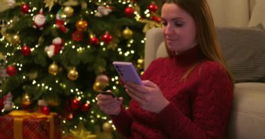 Elinde akıllı telefon ve kredi kartı olan bir kadın süslü Noel ağacının yanında ışık ve süslemelerle oturuyor. Tatil sezonunda online alışveriş. Kapalı mekan sahnesi. Yüksek kalite 4k görüntü