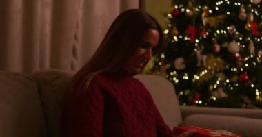 Gülümseyen kadın koltukta otururken parlak kırmızı bir hediye kutusu açıyor. Arka planda ışıklar ve süsler olan Noel ağacı. Memlekette güzel bir tatil akşamı. Yüksek kalite 4k görüntü