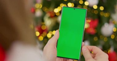 Süslü, bulanık Noel ağacının önünde elinde yeşil ekranlı akıllı telefon tutan bir kadın. Model ve tasarım için yakın çekim. Yüksek kalite 4k görüntü