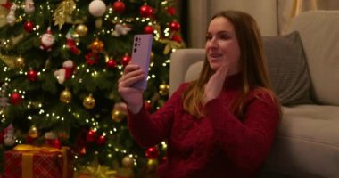 Gülümseyen bir kadın, parlak ışıklarla süslenmiş bir Noel ağacının yanında oturmuş akıllı telefonuyla sohbet ediyor. Yüksek kalite 4k görüntü
