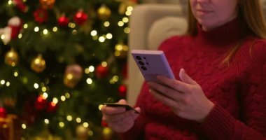 Kırmızı kazaklı bir kadın akıllı telefon ve kredi kartı tutuyor ve ışıkları olan bir Noel ağacının yanında online alışveriş yapıyor. Yüksek kalite 4k görüntü