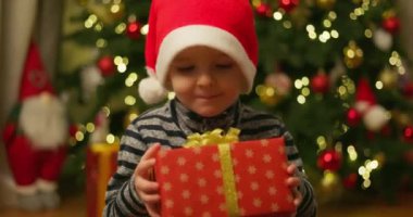 Noel Baba şapkalı bir çocuk, parlak ışıklandırılmış bir Noel ağacının önünde süslerle sarılı bir Noel hediyesi tutuyor. Yüksek kalite 4k görüntü