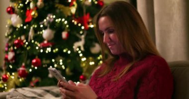 Arka planda süslü Noel ağacı olan bir kadın oturma odasında akıllı telefon kullanıyor. Tatil sezonu ve iletişim konsepti. Sıcak ev atmosferi. Yüksek kalite 4k görüntü
