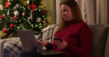 Samimi kırmızı kazaklı bir kadın internetten bilgisayarındaki bir kredi kartıyla Noel ağacının yanında alışveriş yapıyor. Tatil alışverişi ve e-ticaret konsepti. Yüksek kalite 4k görüntü