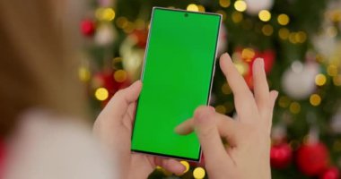 Süslü, bulanık Noel ağacının önünde elinde yeşil ekranlı akıllı telefon tutan bir kadın. Model ve tasarım için yakın çekim. Yüksek kalite 4k görüntü