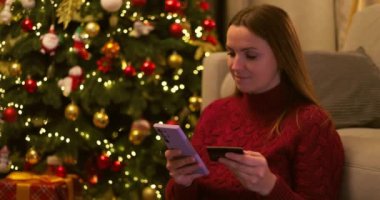 Kırmızı kazaklı bir kadın internetten alışveriş yapmak için akıllı telefon ve kredi kartı kullanıyor, süslü bir Noel ağacının yanında oturuyor. Yüksek kalite 4k görüntü