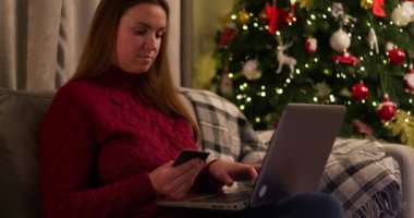 Samimi kırmızı kazaklı bir kadın internetten bilgisayarındaki bir kredi kartıyla Noel ağacının yanında alışveriş yapıyor. Tatil alışverişi ve e-ticaret konsepti. Yüksek kalite 4k görüntü
