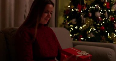 Rahat bir süveter giyen bir kadın, güzel aydınlatılmış bir Noel ağacının yanında parıldayan bir hediye kutusunu açar ve bayram neşesini ve sürprizini yakalar. Yüksek kalite 4k görüntü