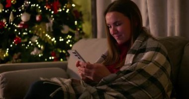 Şenlikli ışıklar ve süslemelerle süslenmiş bir Noel ağacının yanında akıllı telefon kullanırken bir kadın gülümsüyor. kalite 4k görüntü