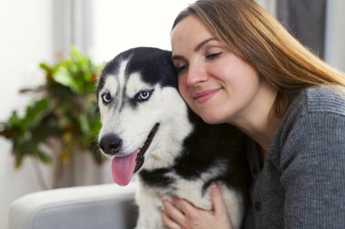 Sibiryalı bir köpeğe evde sarılırken sevgi gösteren bir kadın. Husky mavi gözlü ve mutlu görünüyor, bir kanepede oturuyor. Rahat bir ortamda doğal ışık.