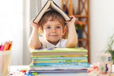 Genç bir çocuk kafasında açık bir kitapla gülümsüyor, masanın üzerindeki renkli kitapların yanında oturuyor. İçerde, arka planda eğitici öğeler bulunan doğal ışık ayarı.