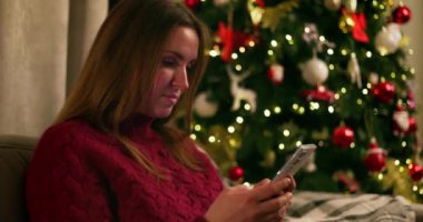 Arka planda süslü Noel ağacı olan bir kadın oturma odasında akıllı telefon kullanıyor. Tatil sezonu ve iletişim konsepti. Sıcak ev atmosferi. Yüksek kalite 4k görüntü