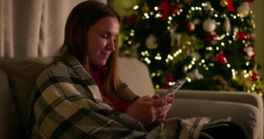 Şenlikli ışıklar ve süslemelerle süslenmiş bir Noel ağacının yanında akıllı telefon kullanırken bir kadın gülümsüyor. kalite 4k görüntü
