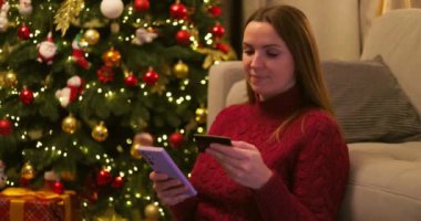 Kırmızı kazaklı bir kadın internetten alışveriş yapmak için akıllı telefon ve kredi kartı kullanıyor, süslü bir Noel ağacının yanında oturuyor. Yüksek kalite 4k görüntü
