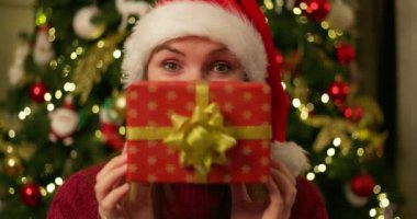 Noel Baba şapkalı bir kadın Noel hediyesini yüzünün önünde süslenmiş bir ağaçla tutuyor. Yüksek kalite 4k görüntü