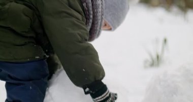Kışlık kıyafetlerle dışarıda kardan adam yapan çocuk. Yeşil ceketli, gri şapkalı ve eşarplı çocuk karda oynuyor. Kış aktivitesi ve tatil konsepti. Yüksek kalite 4k görüntü