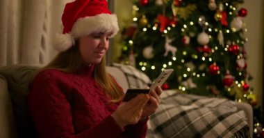 Noel Baba şapkası takan bir kadın online alışveriş için akıllı telefon ve kredi kartı kullanıyor, süslü bir Noel ağacının yanında oturuyor. Yüksek kalite 4k görüntü