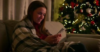 Şenlikli ışıklar ve süslemelerle süslenmiş bir Noel ağacının yanında akıllı telefon kullanırken bir kadın gülümsüyor. kalite 4k görüntü