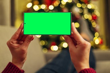 Bulanık bir Noel ağacının önünde yeşil ekranlı akıllı telefonu tutan eller. Aygıt arka planda bokeh ışıkları ile yatay olarak konumlandırıldı.