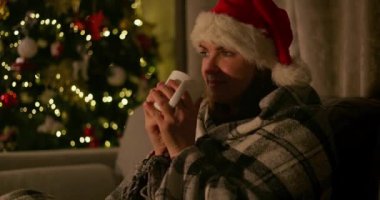 Noel Baba şapkalı ve ekose battaniyeli bir kadın süslü bir Noel ağacının yanında sıcak bir içkinin tadını çıkarıyor ve rahat bir tatil ortamı yaratıyor. Yüksek kalite 4k görüntü