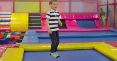 Çizgili tişörtlü genç bir çocuk arka planda köpük çukuruyla kapalı bir oyun merkezinde renkli bir trambolinde zıplıyor. Yüksek kalite 4k görüntü