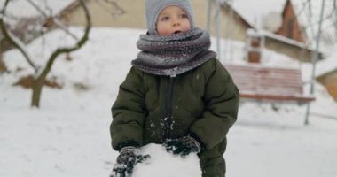 Kışın giyinmiş bir çocuk karlı bir bahçede kartopu yapıyor. Bir yerleşim bölgesinde kış aktivitesi. Çocukluk oyun zamanı ve mevsimsel eğlence konsepti. Yüksek kalite 4k görüntü