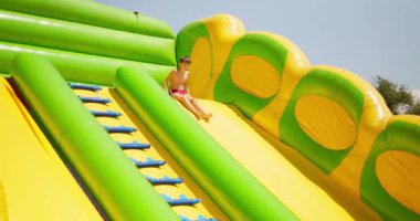 Kırmızı yüzme şortlu genç bir çocuk, güneşli bir günde su parkında canlı yeşil ve sarı bir su kaydırağından kayar. Yüksek kalite 4k görüntü