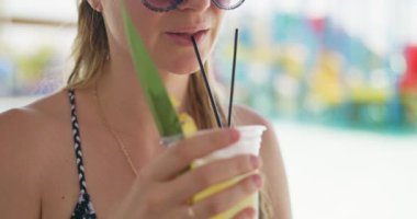 Tropikal bir içeceği siyah pipetle yudumlayan, güneş gözlüğü takan ve elinde ananas yaprağı süslemesiyle içki tutan bir kadının yakın çekimi. Yüksek kalite 4k görüntü