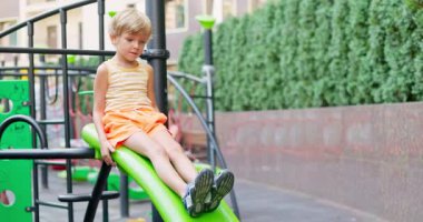 Çizgili askılı tişörtlü ve turuncu şortlu genç bir çocuk yeşil slaytta, çevrenin yeşillikle çevrili olduğu modern bir açık hava oyun alanında oturuyor. Yüksek kalite 4k görüntü