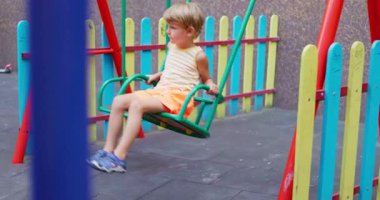 Çizgili askılı tişört giyen ve turuncu şortlu bir çocuk renkli eskrim ve ekipmanlarla dolu canlı bir oyun alanında sallanıyor. Yüksek kalite 4k görüntü