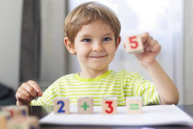 Yeşil çizgili tişörtlü, tahta matematik bloklarıyla oynayan gülümseyen genç bir çocuk ek denklem gösteriyor. Elinde bir numara tutuyor ve bir masada oturuyor. Parlak bir iç mekanda.
