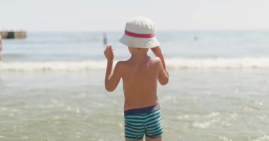 Güneş şapkalı ve yüzme şortlu genç bir çocuk sığ okyanus suyunda, hareketli bir sahilde güneşli bir günün tadını çıkarıyor. Yüksek kalite 4k görüntü