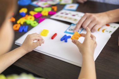 Bir çocuk not defterindeki renkli plastik harflerle yazmayı öğrenirken, bir yetişkin eğitim faaliyet kağıdına işaret eder. Kapalı ev ya da sınıf öğrenme ortamı