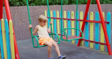 Sarı çizgili tişört ve turuncu şort giyen, renkli çitlerle dolu bir parkta salıncakta sallanan genç sarışın çocuk. Yüksek kalite 4k görüntü