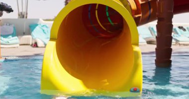 Kırmızı gözlük takan ve çizgili yüzme şortu giyen genç bir çocuk, açık sarı bir su kaydırağından kayarak tatil köyündeki bir havuza kayıyor. Yüksek kalite 4k görüntü