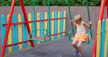 Sarı çizgili tişört ve turuncu şort giyen, renkli çitlerle dolu bir parkta salıncakta sallanan genç sarışın çocuk. Yüksek kalite 4k görüntü