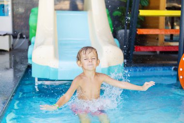 Plastik su kaydırağından yüzme havuzuna atlayan, kolları açık ve su çarpmasına tepki veren tişörtsüz bir çocuk.