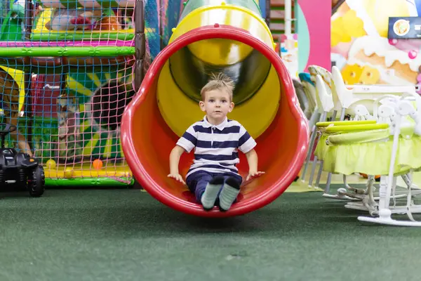 Çizgili tişört giyen genç çocuk kapalı bir oyun bahçesinde kırmızı ve sarı bir tüp kaydırağından kayarak iniyor. Arka planda tırmanma alanları ve oyuncaklar var.