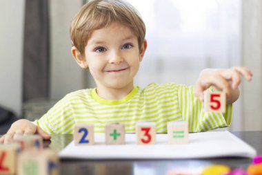 Çizgili gömlek giyen küçük bir çocuk tahta bloklar kullanarak matematik öğreniyor. Çocuk basit bir toplama denklemi oluşturmak için sayıları bir masanın üzerine yerleştirir. Parlak iç mekan