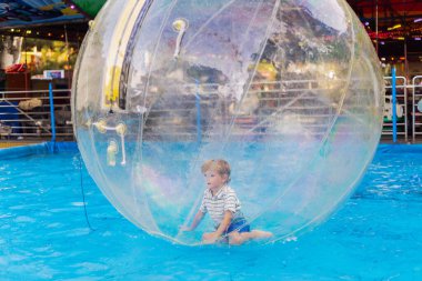 Bir lunaparkta ya da panayırda mavi bir havuzda yüzen şeffaf şişme su topu içindeki çocuk.