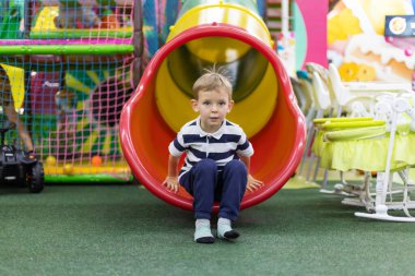 Çizgili tişörtlü çocuk kırmızı ve sarı tüpten çıkıp kapalı alandaki yapay yeşil çimlere kayıyor. Stüdyo yaşam tarzı fotoğrafçılığı. Çocuk aktivitesi ve eğlence konsepti