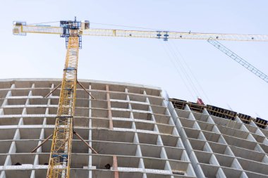 Yapım aşamasındaki çok katlı bir binanın beton iskeleti, açık gökyüzü arka planına karşı çalışan büyük sarı ve mavi vinçler.