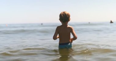 Mutlu çocuk yaz gününün tadını çıkarıyor, açık mavi gökyüzünün altında parlak dalgalarla sığ deniz suyunda duruyor, tasasız ve neşeli. Yüksek kalite 4k görüntü