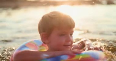 Gökkuşağı şişirilebilir halkasıyla gölde yazın tadını çıkaran, altın saat açık havada yüzerken su sıçrayan ve gülümseyen genç bir çocuk. Yüksek kalite 4k görüntü