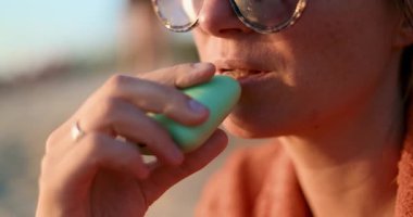 Güneş gözlüğü takıp battaniyeye sarılmış, altın saat boyunca dışarıda pastel yeşil bir tüp kullanan bir kadının yakın çekimi. Yüksek kalite 4k görüntü