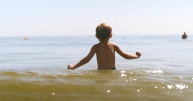 Genç bir çocuk kumsalda güneşli bir günün tadını çıkarıyor. Arka planda insanlar yüzerken sakin deniz sularına dalıyor. Yüksek kalite 4k görüntü