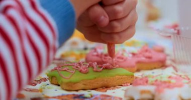 Bir çocuk kurabiyeleri yakın plan çekimde krema ve şekerle süslüyor. Sahne, bayram veya aile temalı içerik için ideal bir şenlik ve yaratıcı pişirme etkinliği yakalıyor.