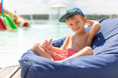 Şapka takan ve çizgili yüzme şortu giyen genç bir çocuk güneşli bir günde havuz kenarındaki mavi fasulye koltukta dinleniyor. Arka planda renkli bir su kaydırağı ve berrak bir gökyüzü var. Neşeli ve yaz havası yaratıyor..