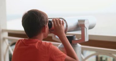 Kırmızı gömlekli genç bir çocuk balkondan manzarayı izlemek için dürbün kullanıyor. Video, onun uzaktaki manzaraya olan merakını ve ilgisini çekiyor..