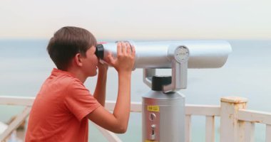 Kırmızı gömlekli genç bir çocuk okyanusu deniz kenarından gözlemlemek için madeni paralı dürbün kullanıyor. Manzaranın tadını çıkarıyor..